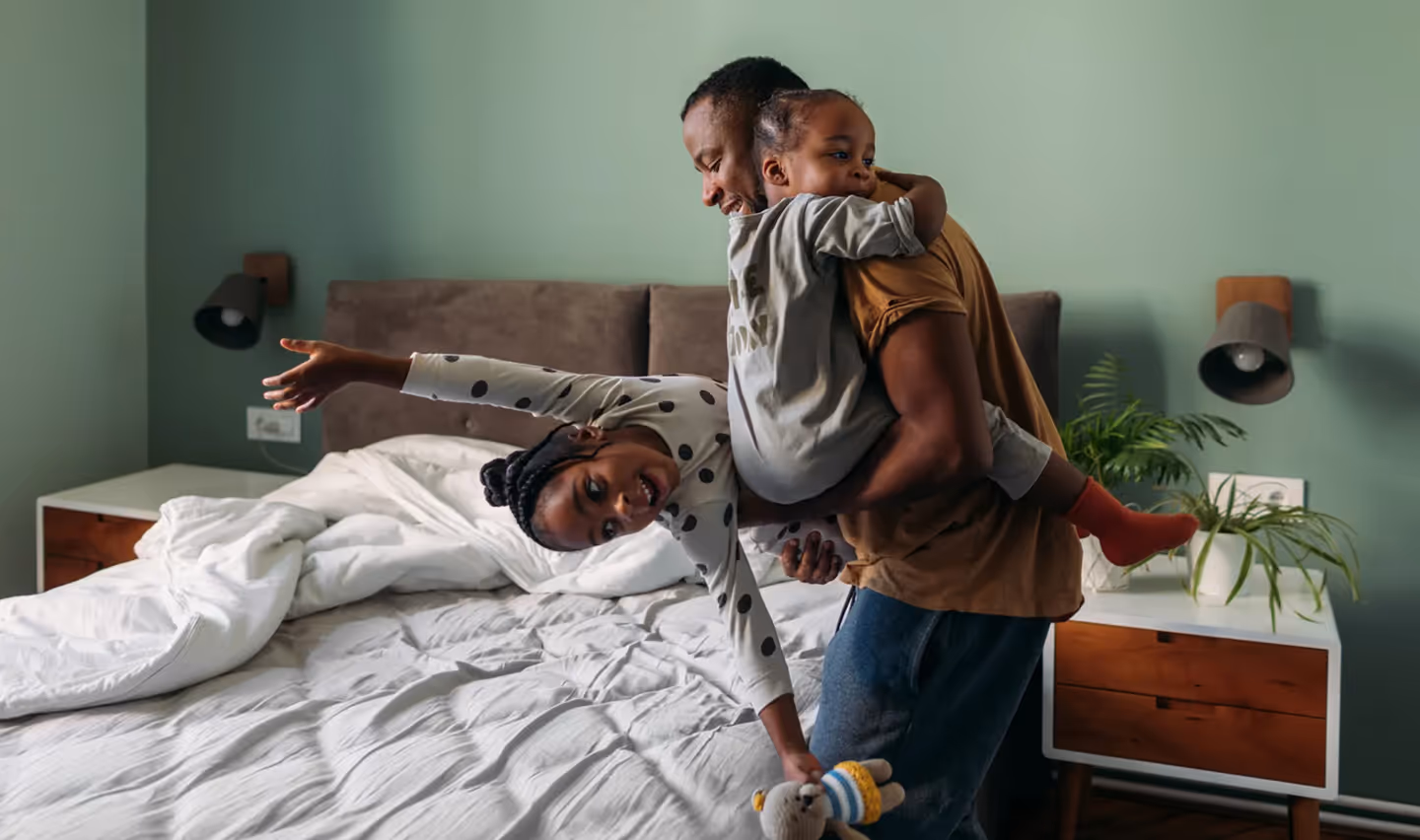 No quarto dos pais, pai brincando com duas filhas e uma delas com braços abertos caindo na cama.